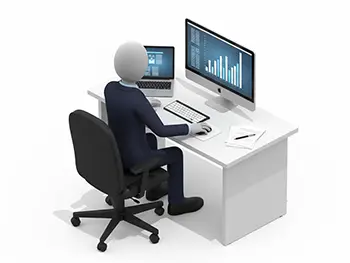 デスクトップパソコンとノートパソコンの画面を見ながらデスクで作業するスーツ姿の人物